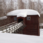 Röd träbyggnad med snötäckt tak och träbro i snöigt landskap med kala träd.
