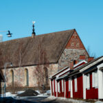 Stenbyggd kyrka med torn och röda träbyggnader längs gata under klar blå himmel.