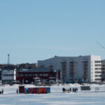Människor promenerar på frusen sjö med snö, byggnader och stor svart kran i bakgrunden.