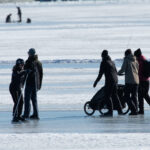 Personer går och åker skridskor på en stor isig och snötäckt sjö, en barnvagn syns.
