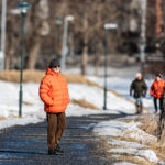 Person i orange vinterjacka står på asfalterad stig med snö och barrträd i vinterlandskap.