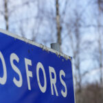 Blå vägskylt med texten "ROSFORS", suddiga träd i bakgrunden.