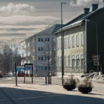 Trottoar och gata med grön träbyggnad och vit flerfamiljsbyggnad, vinter med snö och moln.
