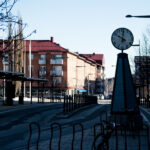 Gata med röda tak, cykelställ, lövfria träd och en stor klocka vid trottoaren.
