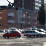 Stadsparkering med flera bilar, grön skåpbil, trädstam och nakna grenar i förgrunden.