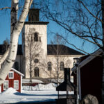 Vinterbild av vit kyrka med torn och röda träbyggnader bland lövfria björkar under blå himmel.