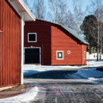 Röda trähus med vita knutar i vintermiljö med snö och lövfria träd under blå himmel.