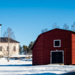 Vit kyrka med svart torn och kors, snötäckt mark och röda träbyggnader under blå himmel.