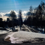 Snötäckt mark med en rak asfalterad väg kantad av lövfria träd under blå himmel med moln.