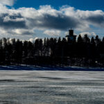 Frusen sjö med skog och en kyrka med kors som silhuett under molnig himmel.