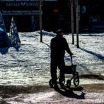 Äldre person med rollator går på isig och snöig yta nära träd och metallskulpturer.