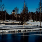 Snötäckt park med frusna vattenytor, lövfria träd och en vit byggnad i bakgrunden.