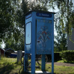 En telefonkiosk med blommigt motiv står i grönska nära byggnad och träd.