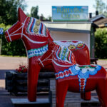 Två röda dalahästar med mönster utomhus, skylt för Rättviks Glass i bakgrunden.