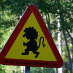 Varningsskylt med silhuett av sagovarelse och webbplats, placerad i skogsmiljö.