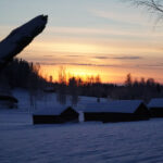 Vinterlandskap vid solnedgång med snöklädda träd och lador i förgrunden, himlen orangegul.