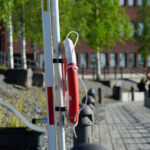 Livboj på stolpe vid stengång i grönområde, joggare och byggnader i bakgrunden.