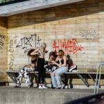 Grupp ungdomar på bänk vid graffitibyggnad, pojke i badbyxor bredvid.