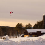 Snölandskap med silobyggnad och person som skärmflyger under molnig himmel.