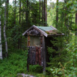 Liten träbyggnad med sadeltak i skog omgiven av gröna träd och buskar.