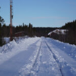 Snötäckt järnväg i skog, klar blå himmel, hus och mast synliga i bakgrunden.