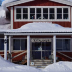 Röd byggnad med snötäckta tak, fönster och veranda med "Kitajaur"-skylt, stege på sidan.