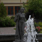 Stenstaty av kvinna vid fontän, byggnad och grönska i bakgrunden.