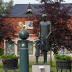Skulptur av vuxen, barn, och gyllene lejon på piedestal utomhus framför tegelbyggnad.
