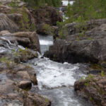 Ström forsar mellan klippor i skog, liten bro i bakgrunden.
