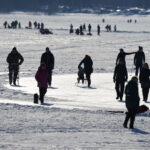 Människor promenerar och sparkar på snötäckt, frusen sjö under solig vinterdag.