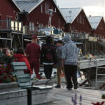 Promenerande människor på brygga vid röda hus, solnedgång, blommor och uteserveringar.