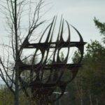 Stor metallskulptur med böjda former i skogsmiljö under mulen himmel.
