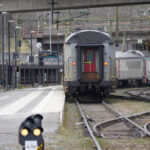 Järnvägsstation med stillastående persontåg, växlar, signaler och bro i bakgrunden.