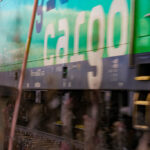 Freight train in motion with side logo, surrounded by tracks and railway infrastructure.