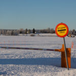 Vinterlandskap med snötäckta fält, träd, bom och "STOПP FÄRJА"-skylt under klar himmel.