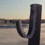 Frosttäckt metallrör i snötäckt fält med skog och solig himmel i bakgrunden.