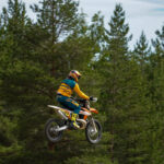 Motorcyklist i färgglada kläder hoppar med motocross i skogsmiljö.
