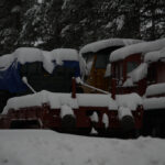 Ett snötäckt tåg står stilla på spår med presenningsklätt fordon, omgiven av snötäckta träd.