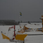 Snötäckt landskap med gul färja, svensk flagga, plogbil och skog i bakgrunden, grå himmel.