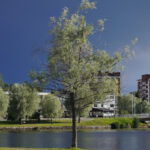 Stadsmiljö med flod, grönska, tegelbyggnader och bro under mörk dramatisk himmel.