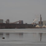 Industribyggnad nära vatten, rök från skorsten, vinter, snö på marken.