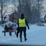 Person i gul reflexväst går på snötäckt väg bredvid gård med röda hus och träd.