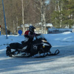 En person på snöskoter på snötäckt väg med träd, byggnader och trafikskyltar i bakgrunden.