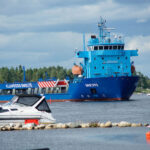 Blått fraktfartyg "KLAIPĖDOS SMELTĖ" seglar på flod, vit motorbåt i förgrunden, skog runtom.