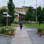 Person går över övergångsställe mot byggnad med skyltar, träd och blommor runt omkring.