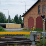 Järnvägsmiljö med tegelbyggnad, stora röda dörrar, grönt tåg och gult skjul.