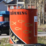 Stor röd cylinder märkt "STRÖMMING" vid vägkorsning med skyltar och en bil i närheten.