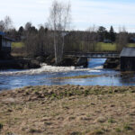 Å rinner förbi äldre byggnader och bro, omgiven av träd och fält i lantlig miljö.