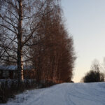 Snötäckt väg med träd och en byggnad till vänster i vinterlandskap.