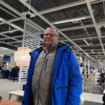 Person in blue jacket and glasses in a store with furniture and people in the background.